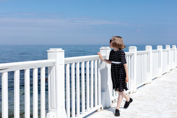 Small (7 years old) pretty cheerful girl in a black  dress with white polka dots is standing on a wooden white pier.