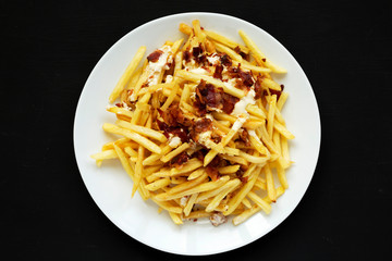 Top view, french fries with cheese sauce and bacon on a white plate over black background. Flat lay, from above, overhead.