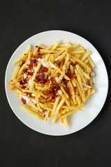 French fries with cheese sauce and bacon on a white plate over black background, top view. Flat lay, from above, overhead.