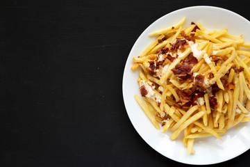 Tasty fast food: french fries with cheese sauce and bacon on a white plate over black background. Flat lay, from above, overhead. Copy space.