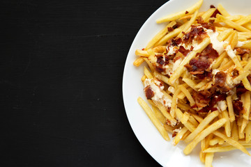 French fries with cheese sauce and bacon on a white plate over black surface, top view. Flat lay, from above, overhead. Copy space.