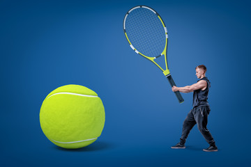 Small man in black sport clothes holding big tennis racket in hands with yellow tennis ball on blue...