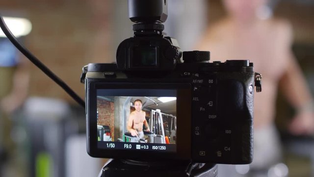 Close-up of camera recording video of shirtless muscular man pointing and saying something before doing leg raise on parallel bars in gym