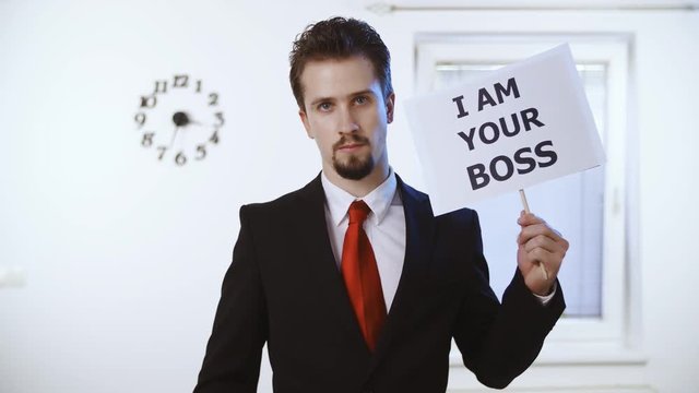 Businessman holds a sign I am your boss 4K. Medium shot of a male person in focus dressed up nicely with a red tie. Wall with clock and window in the background.