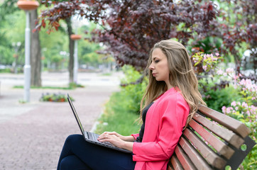 Beautiful young woman using laptop in a park. Portrait of a girl working on computer sitting on the bench on the blossoming street, urban scene