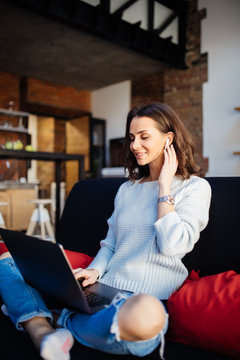 Smiling woman wearing air buds, using laptop at home, sitting on sofa in living room, looking at screen, making video call, working support operator, translator, watching webinar