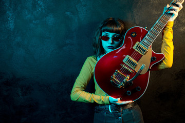 Fashion young hipster woman with curly hair with red guitar in neon lights. Rock musician is...