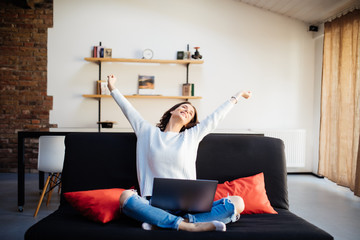 Young woman raising her hands up on the couch with laptop at home