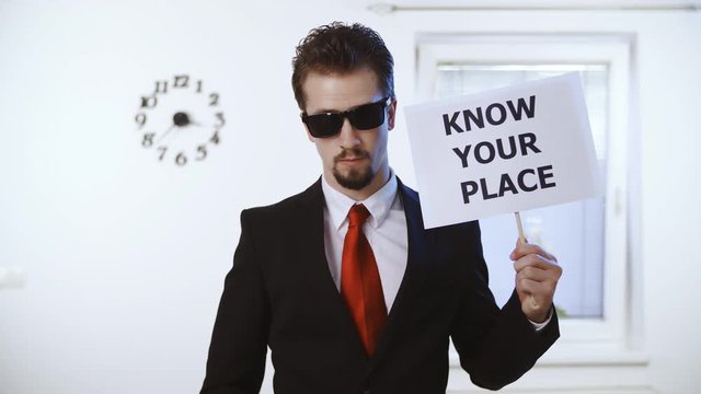Bad ass confident man hold sign Know your place 4K. Medium shot of a male person in focus dressed up nicely with a red tie. Wall with clock and window in the background.