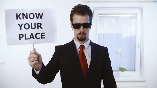 Businessman hold a sign Know your place 4K. Medium shot of a male person in focus dressed up nicely with a red tie. Wall with clock and window in the background.