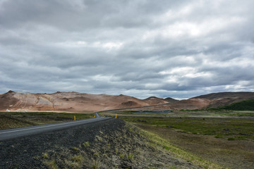 Curvy road, empty meadow and red mountains in the background in Myvatn region, overcast day in summer