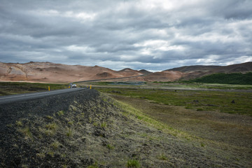 Fototapeta na wymiar Road, empty meadow and red mountains in the background with a car in Myvatn region, overcast day in summer