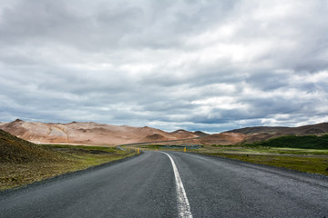 View to the empty curvy road, empty meadow and red mountains in the background in Myvatn region, overcast day in summer