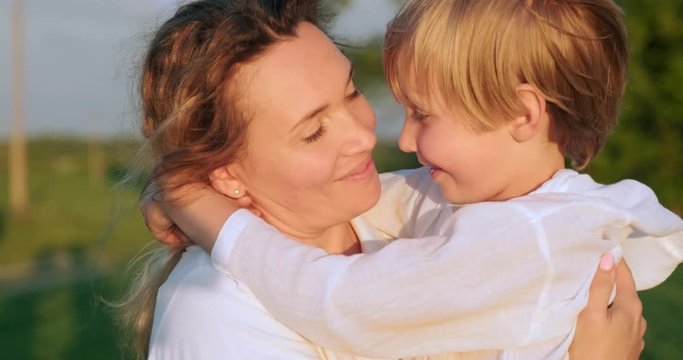 Happy mother  kissing her son on hands walking on the meadow in summer.  Mother with 7 year child strolls on field.   Adult mother with blond boy sauntering in nature. Slow motion. Happiness.