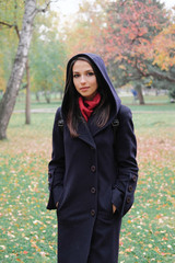 The girl in the black coat looks away from the camera on background of autumn trees.