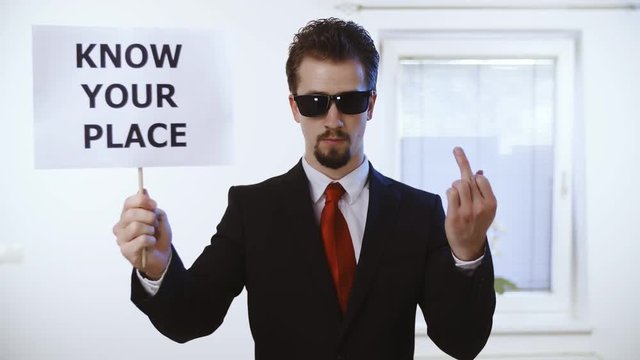 Man in suit hold a sign Know your place with middle finger 4K. Medium shot of a male person in focus dressed up nicely with a red tie. Wall with clock and window in the background.
