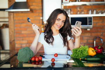 Young happy woman holding spoon and choose recipe with eco vegetables, look into web the phone and sitting in the kitchen