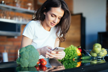 Smiling young woman text messaging in front of vegetables in the kitchen at home