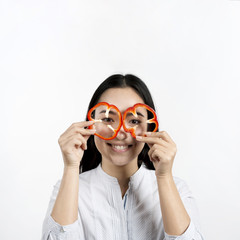 Asian woman holding two slices of red bell peppers in front of her eyes above white surface