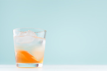 Summer striped oranges lemonade with ice cube, liquor in misted glass on elegant pastel green wall, white wood table.