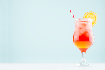 Sunrise layered red, yellow drinks with orange, straw, ice cubes in elegant glass on pastel blue...