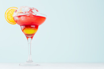 Summer fresh fruit layered red, yellow sunrise cocktail with orange slice, ice cubes in wineglass on pastel green background.
