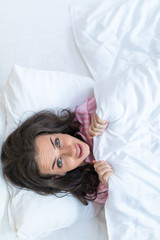 Fototapeta premium Girl hides her face in bed. Cheerful girl in bed hiding her face under the blanket. Teenage girl lying under a blanket. Woman playful with a blanket view from above. Girl peeks out from the duvet.