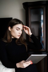 Beautiful young woman in casual clothes is using a digital tablet and smiling while sitting on couch at home