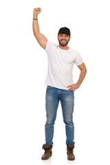 Fototapeta premium Young Man In Jeans, White T-shirt And Black Cap Is Standing With Arm Raised And Smiling