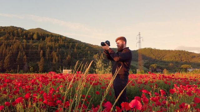 Giovane fotografo con barba lunga e vestito di nero sta scattando delle fotografie in mezzo a un campo di papaveri rossi al tramonto.