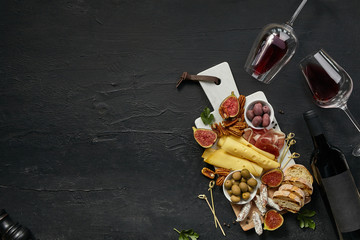 Obraz na płótnie Canvas Two glasses of red wine and a tasty cheese plate with fruit, grape, nuts and toasted bread on a wooden kitchen plate on the black stone background, top view, copy space. Gourmet food and drink.
