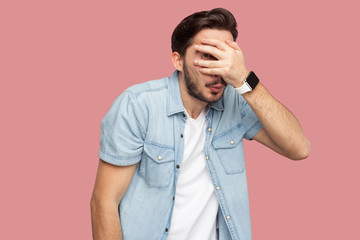 Portrait of shy, scared or peeking bearded young man in blue casual style shirt standing covering his eyes and looking at camera through his fingers. indoor studio shot, isolated on pink background.