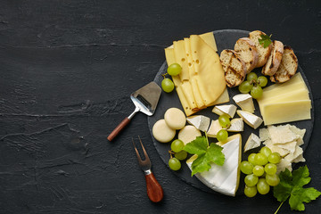 Top view of tasty cheese plate with fruit, grape on a circle kitchen plate on the black stone background, top view, copy space. Gourmet food and drink.