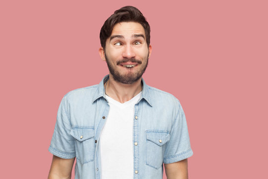 Portrait of crazy handsome bearded young man in blue casual style shirt standing with crossed eyes and looking with funny comedian face. indoor studio shot, isolated on pink background.