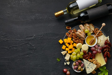 Top view of tasty cheese plate and wine bottles with fruit, grape, nuts and honey on a circle kitchen plate on the black stone background, top view, copy space. Gourmet food and drink.