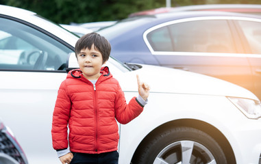 Outdoor portrait preschool kid boy with a funny face standing next to the car, Happy child pointing making fun face and showing finger up standing alone in car park