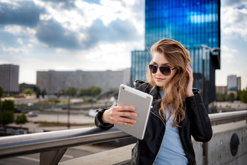 Fototapeta premium Young smiling woman using tablet among modern office buildings on city street
