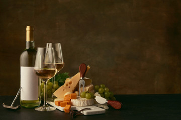 Fototapeta na wymiar Front view of tasty cheese plate with grapes and the wine bottle, fruit and wineglasses on dark studio background, copy space to insert your text or image. Gourmet food and drink.