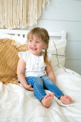 Fototapeta premium Beautiful little girl sitting on the bed in the bedroom. the baby has blonde hair and blue eyes. Jeans and white t-shirt