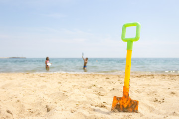 Yellow plastic toy shovel on sand beach at the seaside and kids playing in the sea in background.