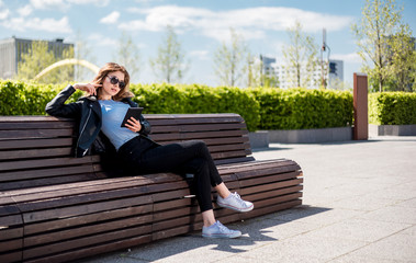 Fototapeta premium Woman sitting on bench among urban space and using tablet in the city