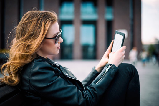 Woman sitting on bench among urban space and reading ebook using digital reader