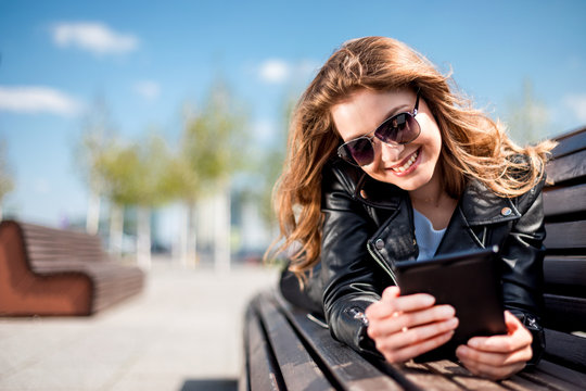 Woman sitting on bench among urban space and reading ebook using digital reader
