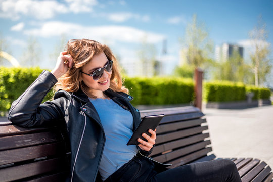 Woman sitting on bench among urban space and reading ebook using digital reader