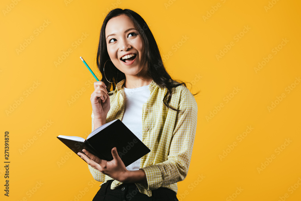 Wall mural smiling asian girl with notebook and pen, isolated on yellow