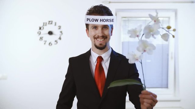 Naive rich businessman plow horse 4K. Medium shot of a male person in focus dressed up nicely with a red tie. Wall with clock and window in the background.