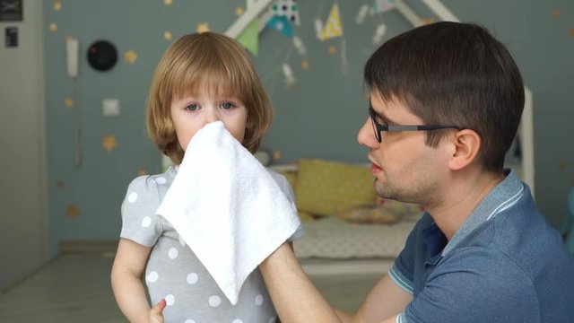 Portrait of young father with glasses wipes runny nose and tears of little cute daughter with napkin. Baby girl cries because of illness and snot