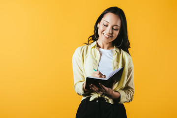 attractive asian girl writing in notebook with pen, isolated on yellow