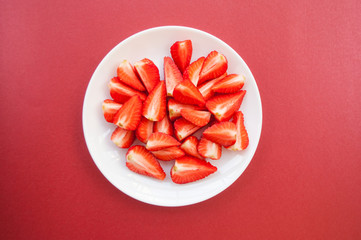 Fresh juicy cut strawberries on a white plate on dark red background with copyspace. Flat lay style.