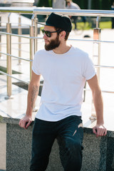 Hipster handsome male model with beard wearing white blank t-shirt and a baseball cap with space for your logo or design in casual urban style